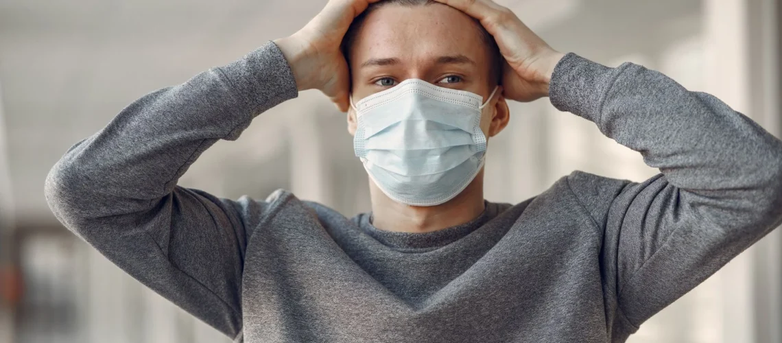 Jeune homme portant un masque médical, la tête entre les mains, exprimant anxiété et stress liés à la pandémie.