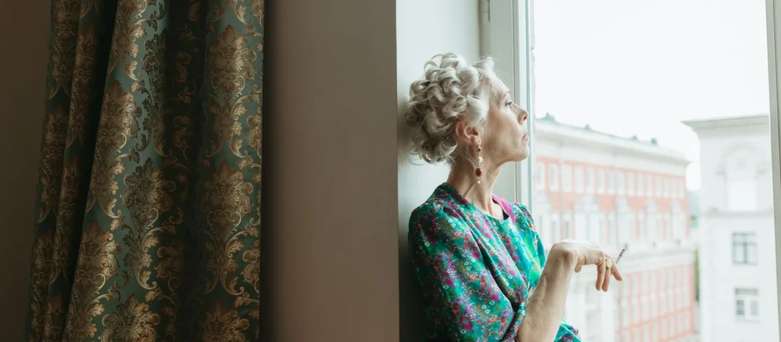 Femme âgée en robe à fleurs fumant près d'une fenêtre à l'intérieur