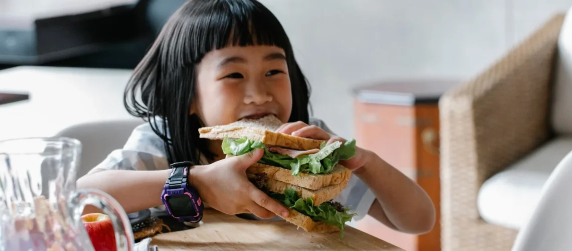 Enfant asiatique assis à une table en bois au petit-déjeuner, mangeant un sandwich garni de laitue.
