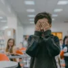 Jeune garçon se couvrant le visage dans une classe bruyante, exprimant stress ou anxiété.