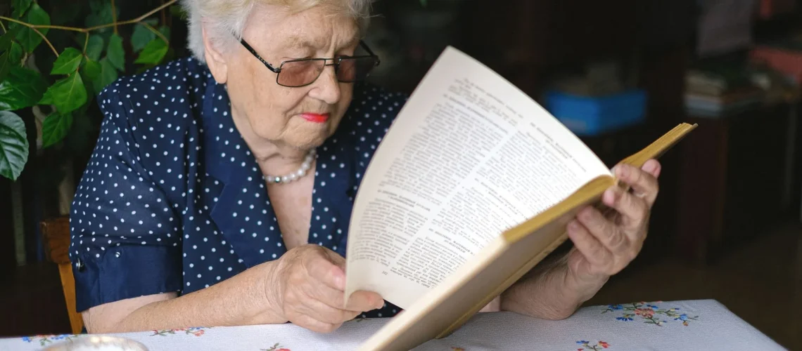 Retraitée sérieuse en lunettes lisant un livre à table avec une boisson chaude, arrière-plan flou.