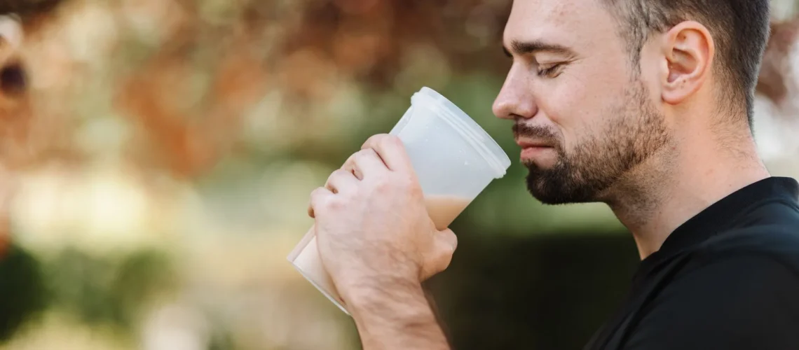 Homme adulte dégustant un shake protéiné dans un cadre extérieur paisible en journée.