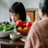 Adorable petit enfant asiatique boudeur mangeant des pommes et de la laitue verte saine lors d'un déjeuner avec sa mère non identifiée dans une cuisine confortable.