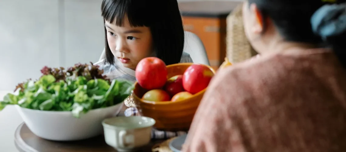 Adorable petit enfant asiatique boudeur mangeant des pommes et de la laitue verte saine lors d'un déjeuner avec sa mère non identifiée dans une cuisine confortable.