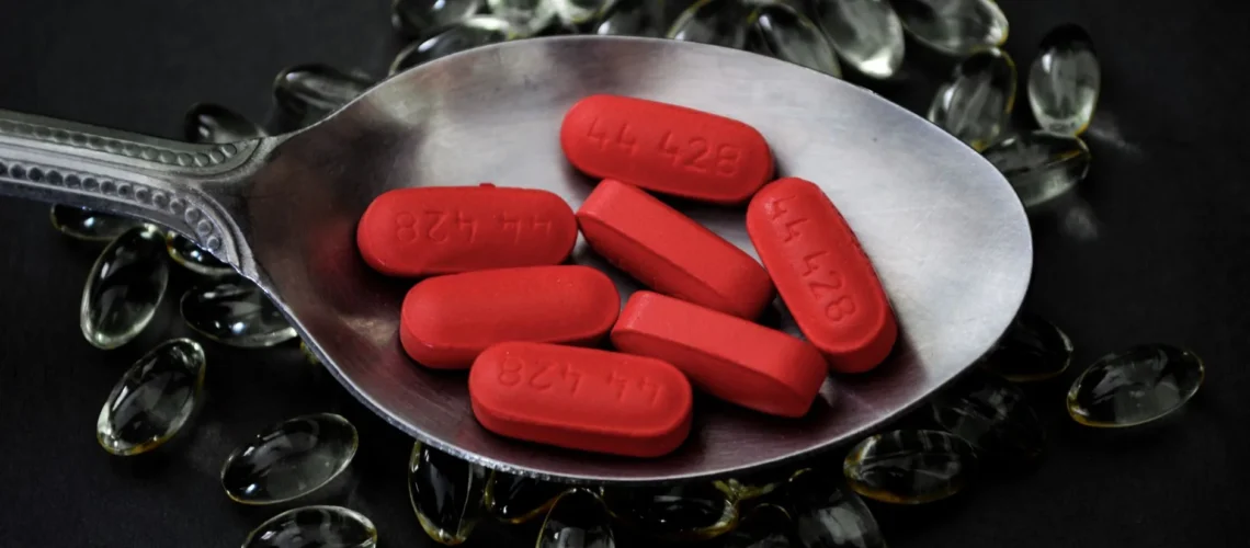 Gros plan sur des comprimés rouges posés sur une cuillère, entourés de gélules transparentes.