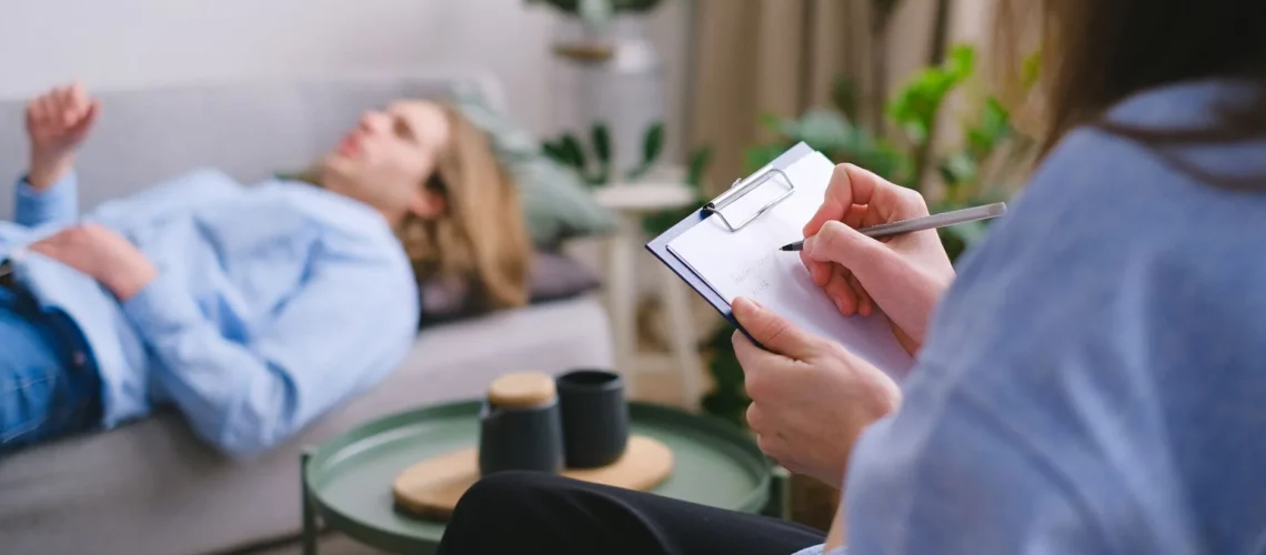 Thérapeute professionnel non identifiable écrivant sur un porte-bloc, assis face à un client masculin allongé sur un canapé, arrière-plan flou, lors d'une thérapie de santé mentale.