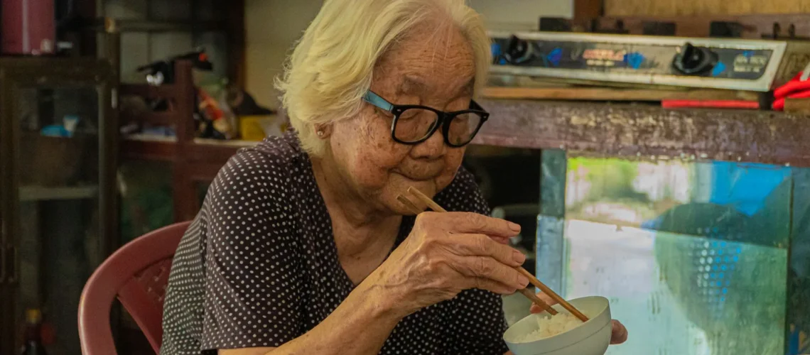 Femme asiatique âgée mangeant du riz avec des baguettes dans un cadre intérieur confortable.