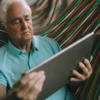 Homme âgé se détendant dans un hamac tout en utilisant une tablette, vêtu d'un polo bleu clair
