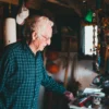 Un homme âgé en chemise à carreaux explore un espace intérieur encombré d'objets personnels et d'un drapeau canadien.