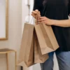 Une personne debout à l'intérieur tient trois sacs en papier brun dans une main, en tenue décontractée.